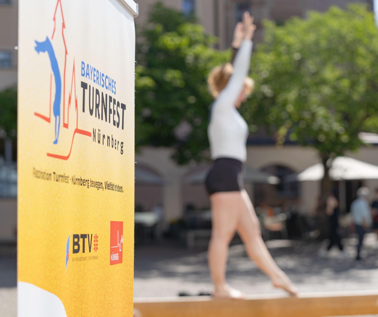 Banner des Bayerischen Turnfests Nürnberg, im Hintergrund turnende Person auf dem Schwebebalken auf einem Platz. Foto: Claudia Doenitz / Bayerischer Turnverband e. V.