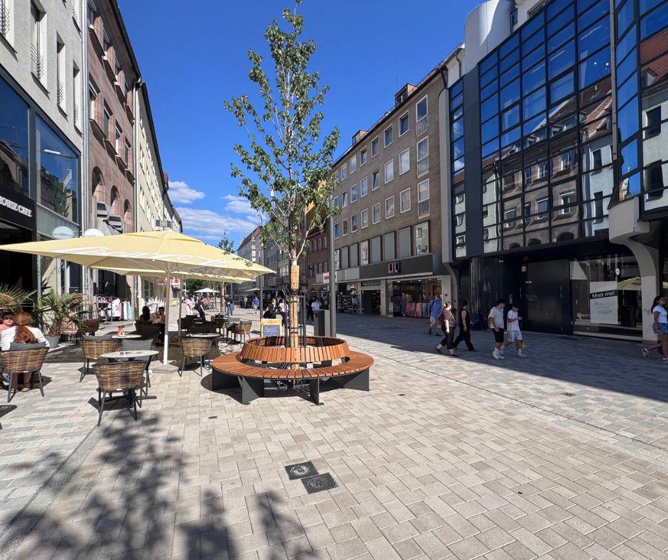 Perspektive vom Weißen Turm in die Breite Gasse zeigt glattes Granitpflaster, gelbe Sonnenschirme, einen jungen Baum mit runder Sitzfläche drumherum, Häuserfronten sowie Menschen, die im Schatten durchs Bild laufen.
